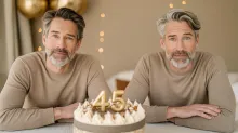 Two middle-aged men with different signs of aging sit behind a birthday cake with golden "45" candles, illustrating the concept of biological age.