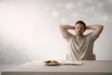 Man sitting at a table with food and notebook, stretching with hands behind head, looking thoughtfully at the camera