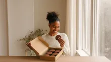 A smiling woman opens a box of cookies and reaches for one, illustrating how product design, convenience, and eating behavior may influence food choices beyond ingredients alone.