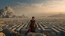 Ancient Greek hero standing before a vast stone labyrinth stretching across a mountainous landscape at sunset.