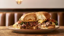 Two halves of a broken cheeseburger on a plate, revealing the soft bun, beef patty, melted cheese, tomato, and lettuce in a warm restaurant setting.