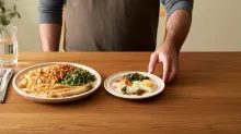 A man stands behind a wooden table with a large plate of food and a smaller nutrient-dense plate containing fish, egg, and vegetables, reaching toward the smaller portion.